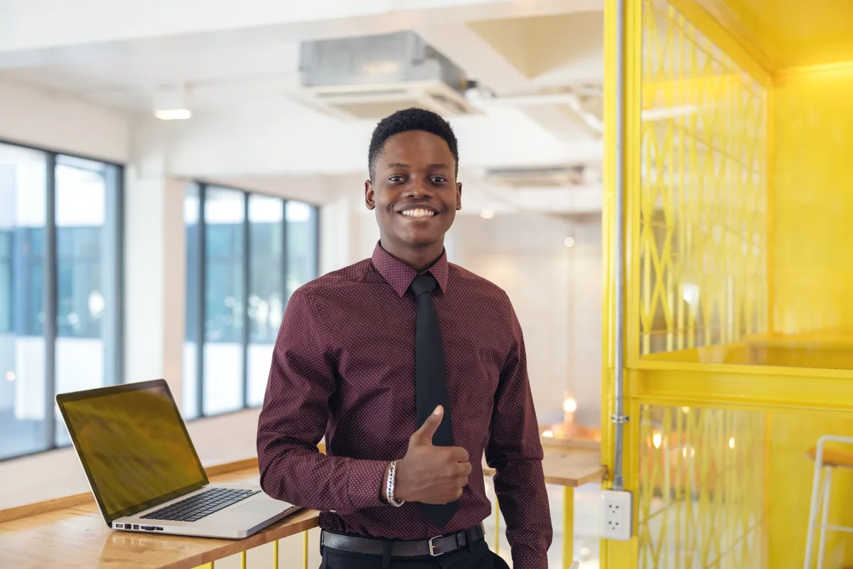 A smiling young professional giving a thumbs up beside his laptop